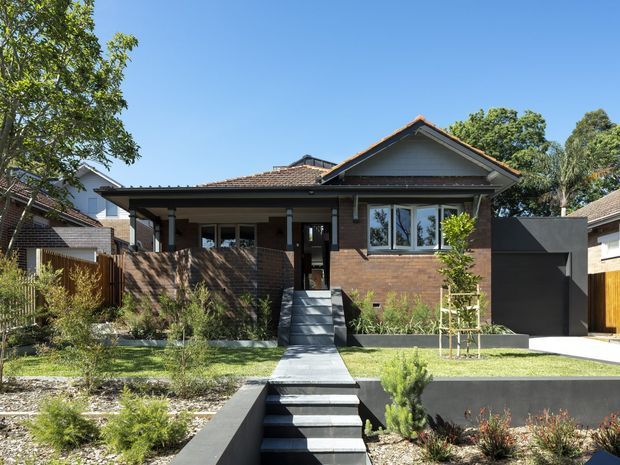 Front exterior view of a suburban single-storey house with a prominent gabled roof, dark brick and timber-toned facade, contrasting dark garage volume, and a landscaped front garden. A stepped concrete path leads up to the recessed entry, with neighbouring homes visible at either side.