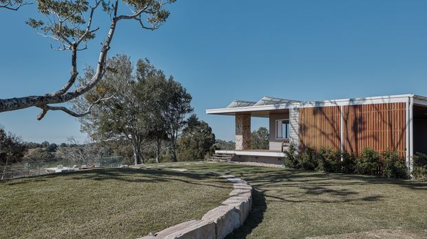 A low, modern single-storey house set within a landscaped grassy site. The building has a broad flat roof with raised glazed roof elements, deep overhanging eaves, large openings, and a prominent facade of vertical timber battens. A curving stone-edged pathway leads across the lawn toward the entrance, with mature trees and an open rural-looking landscape beyond.