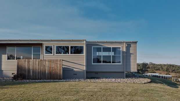 Single-storey contemporary residential dwelling viewed from the exterior, with horizontal weatherboard cladding, a low-pitched roof, and a long glazed facade. A timber privacy fence encloses a small patio or entry area, while a broad manicured lawn and landscaped gravel edging surround the house. The setting appears elevated or sloped, with distant vegetation and a body of water or roadway visible beyond, though these background elements are secondary.