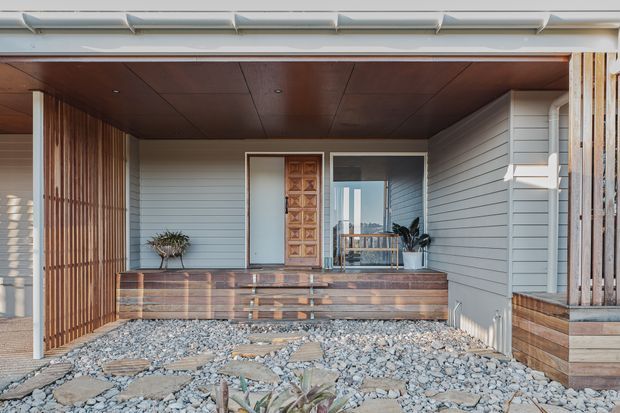 Front entry porch of a contemporary coastal-style home, featuring pale horizontal weatherboard cladding, a warm timber soffit, vertical timber screening, a timber entry door, raised timber-clad planter or bench structures, and a rough stone gravel path with stepping stones. The sheltered recessed entry opens toward a glazed door or window reflecting the surrounding setting.