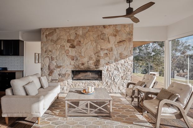 A bright mid-century-inspired living room centred on a large rough stone feature wall with an inset fireplace. The room includes a light upholstered sofa, timber-framed lounge chairs, a geometric coffee table, a stone-tiled floor, expansive glazing to the garden, and a sculptural timber-blade ceiling fan.
