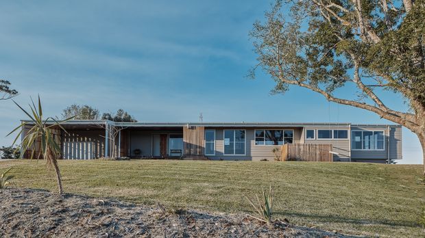 A single-storey, low-profile residential house viewed from across a gently sloping lawn. The long horizontal facade combines light grey weatherboard cladding, timber-toned sections, extensive windows, and a broad covered carport or verandah at the left. Mature trees and sparse landscaping surround the property, with a clear blue sky behind it.
