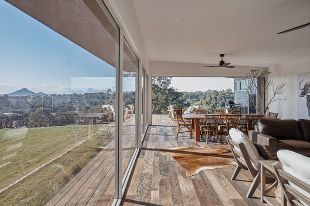 Bright, open-plan living and dining area with extensive floor-to-ceiling glazing overlooking a landscaped rural or semi-rural valley. The room features timber flooring, a timber dining table and chairs, a dark sofa, contemporary ceiling fan, and a spacious timber deck extending outdoors. Natural light and the wide view create an airy, relaxed atmosphere.
