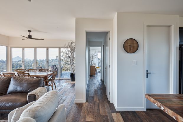A bright contemporary open-plan living and dining area viewed toward a central hallway. The room features large windows with expansive views, a leather sectional sofa, timber dining furniture, wide-plank timber flooring, white walls, minimalist flush doors, and a ceiling fan. A doorway reveals additional interior space beyond.