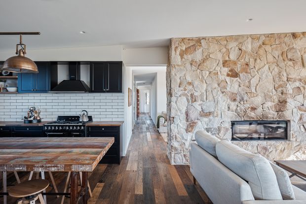 Open-plan interior showing a dark fitted kitchen on the left, a timber dining island in the foreground, and a living area with a large irregular natural-stone feature wall and built-in fireplace on the right. A hallway extends through the centre of the home. The space combines rustic materials with contemporary cabinetry and fittings.