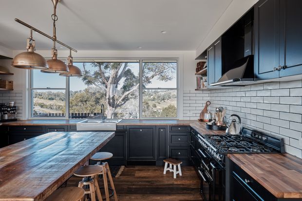A spacious, well-appointed kitchen with dark painted cabinetry, white subway-tile backsplash, timber benchtops and a large timber island with stools. A substantial stainless-steel range cooker sits beneath a black rangehood, while copper pendant lights hang over the island. Large windows provide views to a leafy, hilly landscape, and dark timber flooring adds warmth.