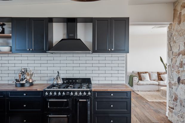 A traditional-style kitchen with dark painted shaker cabinetry, a large black range cooker and hood, and a white horizontal subway-tile backsplash. The timber benchtop adds warmth, while an opening reveals an adjacent sitting area with a light sofa and timber flooring. A rustic stone-clad wall frames the right side of the image.