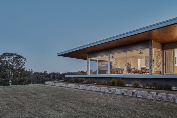 A low, single-storey modern house set in a landscaped rural or bush setting. The image focuses on a broad cantilevered flat roof with timber soffit, expansive floor-to-ceiling glazing, and a stone-clad retaining wall. A furnished open-plan interior is visible through the glass, including seating and dining areas, while the foreground is a large lawn.