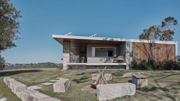 A single-storey contemporary pavilion-style house set within a landscaped grassy site. The home features a broad low-pitched roof with deep eaves, exposed timber soffit, stone-clad structural elements, horizontal wall cladding, and a screened section of vertical timber battens. A covered outdoor living area opens toward a rough stone seating and fire-pit arrangement in the foreground, with mature trees and a distant landscape beyond.