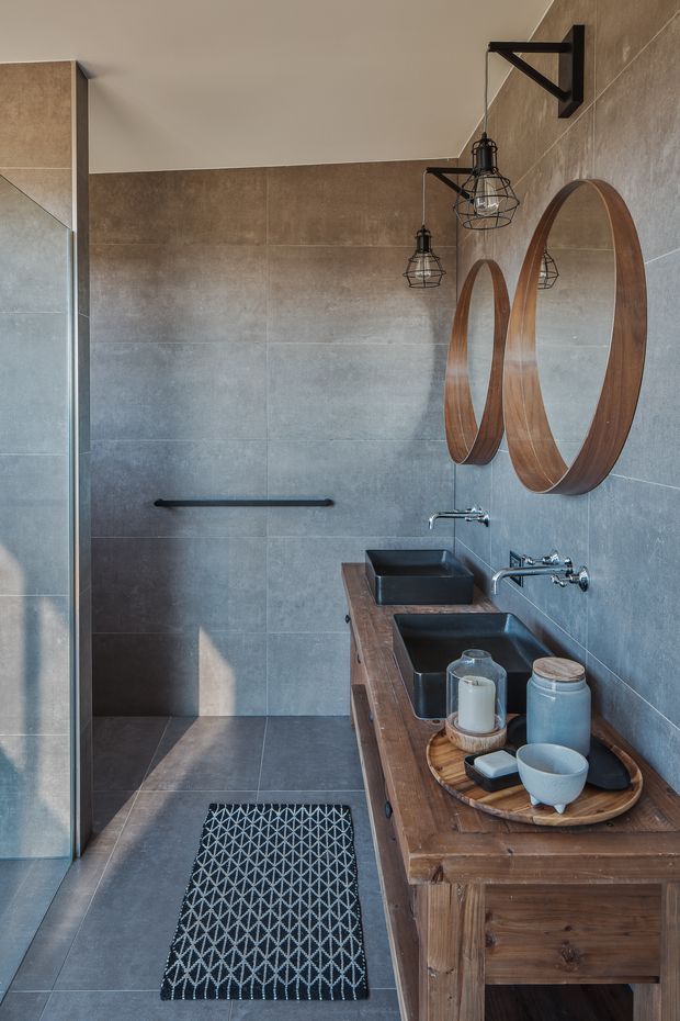 A contemporary bathroom with a walk-in shower, full-height grey-brown stone-look wall and floor tiles, and a rustic timber double vanity. The vanity has three dark rectangular vessel basins with exposed chrome wall-mounted tapware. Large oval timber-framed mirrors and black cage pendant lights add industrial character, while a patterned runner and small countertop accessories provide contrast.