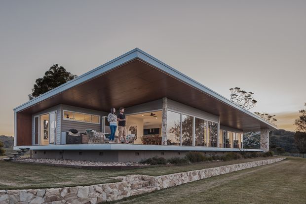 Single-storey contemporary pavilion-style house set in a landscaped rural or bush setting. The home has a broad low-pitched roof with deep cantilevered eaves, extensive floor-to-ceiling glazing, a timber-lined soffit, slender dark window frames, and a raised deck wrapping around an open-plan living area. Natural stone retaining walls and muted landscaping surround the building.