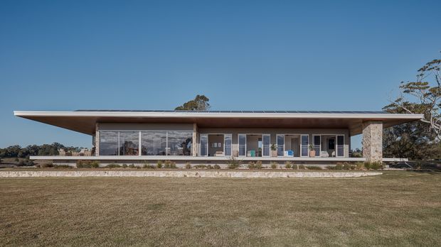 A low, single-storey modern residence viewed from across a broad lawn. The house has a wide shallow roof with deep overhanging eaves, extensive floor-to-ceiling glazing, a long covered terrace, and a prominent stone-clad structural pier at the right. The setting appears spacious and landscaped, with a restrained horizontal composition and clear blue sky.