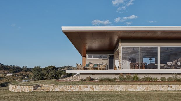 A low-slung contemporary single-storey house viewed from the exterior, set on a landscaped sloping site. The home features broad floor-to-ceiling glazing, a wide projecting flat roof with a timber-lined soffit, pale stone wall cladding and retaining walls, and an outdoor terrace with dining furniture. The surrounding rural landscape and blue sky are visible beyond the building.