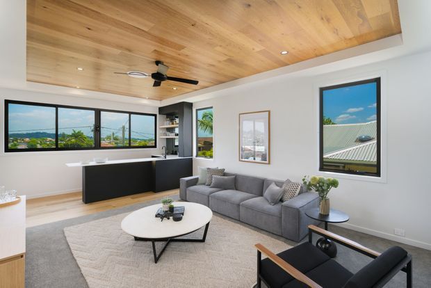 Bright contemporary open-plan living area with a grey sectional sofa, round coffee table, black accent armchair, and large rug. A compact black kitchen with a peninsula sits at the rear, while expansive black-framed windows provide views of greenery and neighbouring rooftops. The room features a warm timber-lined recessed ceiling with a ceiling fan and recessed downlights, complemented by white walls and light flooring.