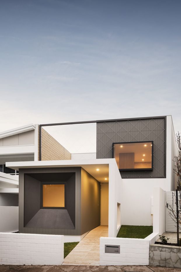 Street-facing view of a compact contemporary townhouse facade with strong geometric forms. The exterior combines white rendered and painted brick walls, dark grey cladding, a recessed timber entry, projecting flat-roof canopy, small lawn areas, and a large upper window with warm interior lighting. A neighbouring dwelling is partially visible at the left.
