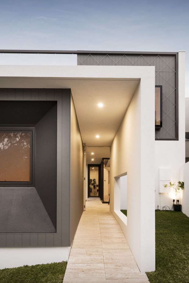 Contemporary residential facade with a long sheltered entry passage, pale tiled paving, crisp white rendered walls, dark grey vertical cladding, and a patterned upper screen. Recessed ceiling lights illuminate the walkway, with a small landscaped lawn and minimalist planting beside the entrance.