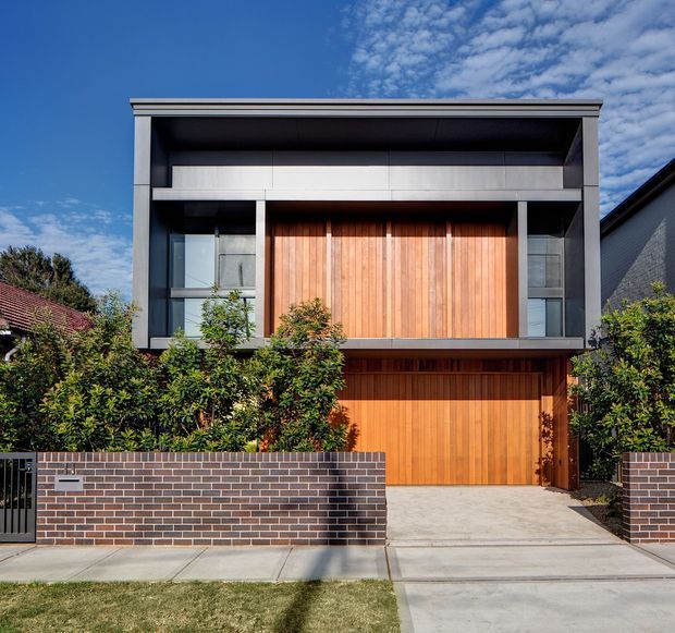 Street-facing view of a contemporary two-storey residence with a flat roof, strong rectangular geometry, dark metal or painted framing, large upper-level glazing, and a prominent vertical timber-clad facade panel. The lower level features a timber garage door, concrete driveway, landscaped screening, and a low dark brick boundary wall.
