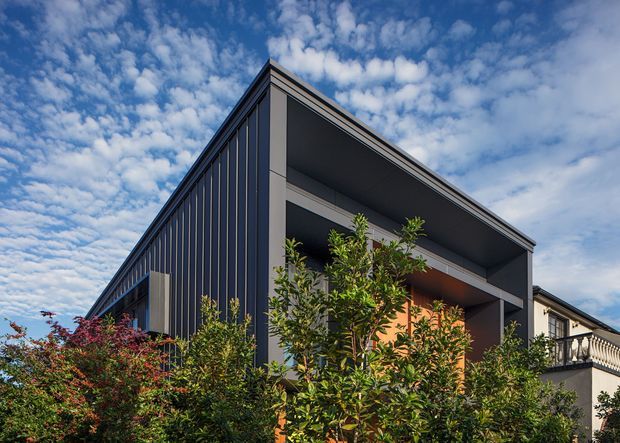 Exterior architectural photograph of a contemporary multi-level residence, dominated by a dark vertically clad side wall and a projecting concrete or rendered balcony volume with deep recessed openings. Warm timber-toned surfaces are visible within the recesses, with landscaping in the foreground and a traditional neighbouring structure at right beneath a partly cloudy sky.