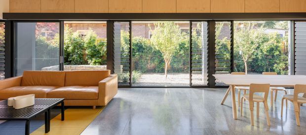 Open-plan contemporary living and dining area with a tan leather sofa, black slatted coffee table, pale wood dining table and chairs, polished grey concrete floor, and extensive black-framed glazing opening views to a leafy garden. Timber wall panelling spans the upper portion of the room, creating a warm contrast with the dark frames and concrete floor.