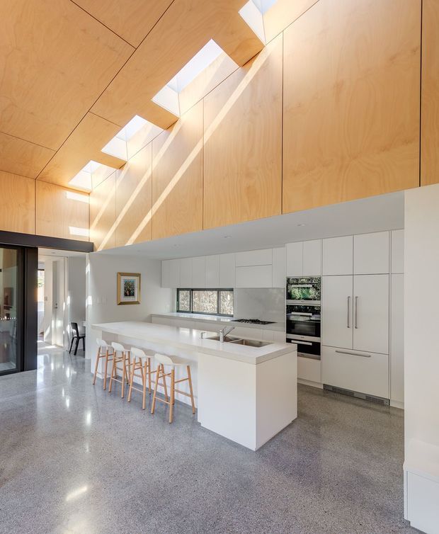 Bright contemporary kitchen with a large white island and integrated seating, extensive white handleless cabinetry, stainless-steel appliances, and polished grey terrazzo flooring. The room features a dramatic warm timber-lined pitched ceiling with a series of skylights, creating a spacious, light-filled interior. Glazed doors and an adjacent dining area are visible at the left.