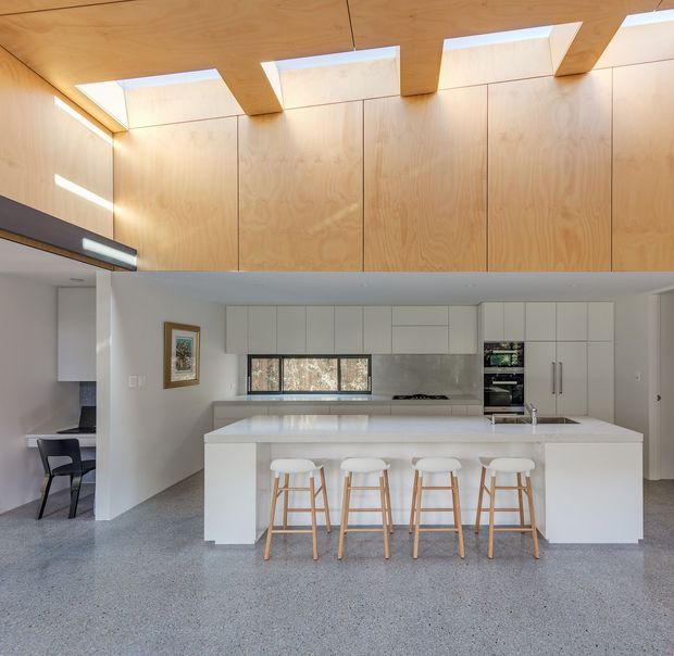 A bright contemporary kitchen with a large white island and four timber bar stools. The room features extensive pale timber wall and ceiling lining, white flat-panel cabinetry, integrated appliances, a stone-look benchtop and splashback, and a polished grey speckled floor. Clerestory-style roof openings bring strong natural light into the space, while a dining area is partially visible to the left.