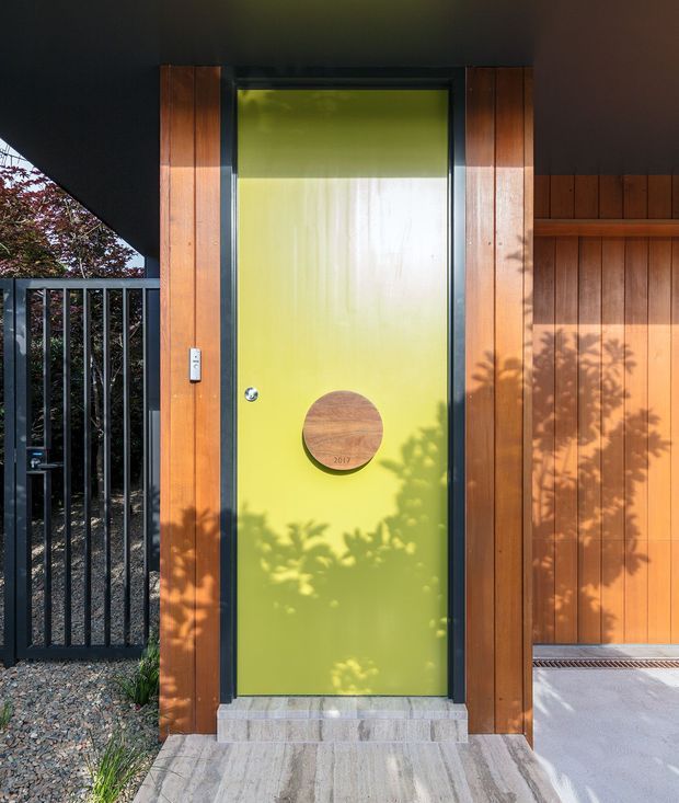 Close-up exterior view of a contemporary entry door set within warm vertical timber cladding. The door is a vivid lime green with a round timber feature panel, dark trim, and a small metal handle. A black metal gate, gravel landscaping, concrete paving, and partial roof overhang frame the entry.