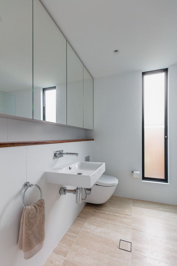 Bright, compact contemporary bathroom with a wall-mounted white rectangular basin, wall-hung toilet, large mirrored cabinetry, slim dark-framed window, and pale wood-look tiled flooring. The room has white tiled walls, a narrow timber shelf, chrome tapware, and a clean minimalist fit-out.