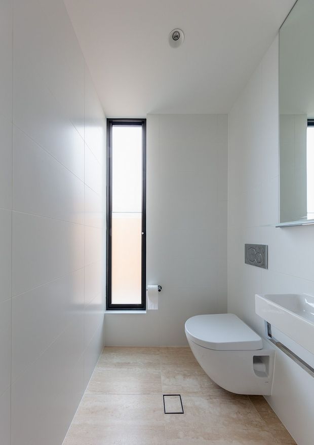 Bright, minimalist contemporary bathroom with a wall-hung toilet, floating white vanity, large mirror, narrow black-framed window, pale tiled walls, and beige stone-look floor tiles. The compact room has clean lines, a flush wall plate, recessed ceiling light, and a linear floor drain.