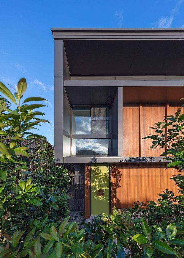 A contemporary multi-storey residential facade viewed from a lush garden. The house features a strong dark grey rectangular frame, deep roof overhangs, large windows, warm vertical timber cladding, and a contrasting green entry door. Dense tropical planting partially screens the lower facade, with bright blue sky visible above.