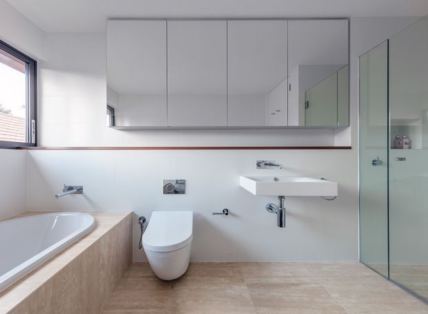 Bright contemporary bathroom with a freestanding bathtub set into a timber-look surround, wall-hung toilet, floating white vanity basin, large mirrored wall cabinetry, and a frameless glass shower enclosure. The room has pale neutral walls, light wood-effect surfaces, chrome fixtures, and a clean minimalist layout.