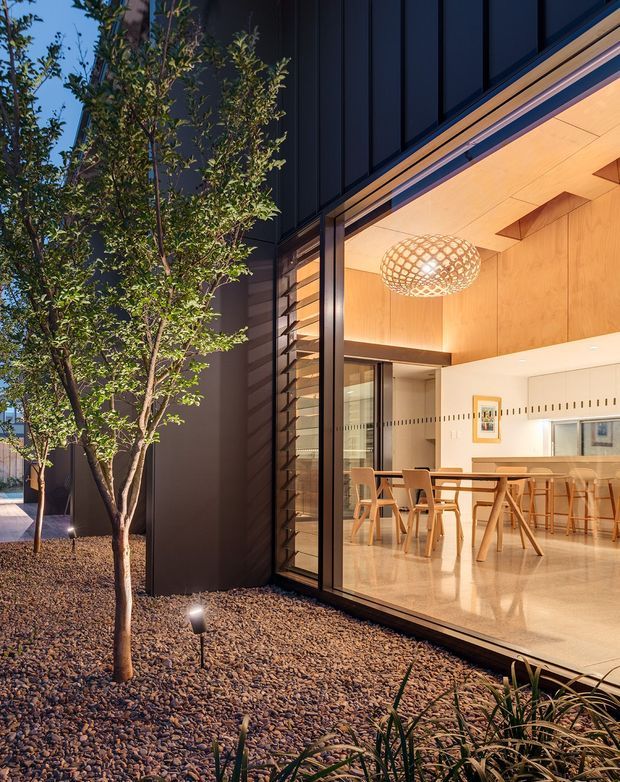 A contemporary house exterior at dusk, viewed from a landscaped gravel courtyard. Large floor-to-ceiling glazing reveals a warm open-plan dining and kitchen area with timber cabinetry or wall lining, a timber dining table and chairs, and a sculptural woven pendant light. The exterior is clad in dark vertical panels with timber-toned soffits and reveals, while small trees and low planting occupy the rock-covered garden.