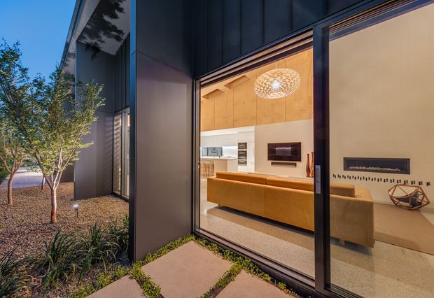Twilight view of a contemporary home's living room through expansive floor-to-ceiling glazing. The interior features warm timber wall and ceiling lining, a long mustard-brown sofa, pale walls, polished concrete or terrazzo-like flooring, minimalist built-in cabinetry, and a sculptural pendant light. Dark cladding and slender dark window frames contrast with the warmly lit interior, while a planted landscaped garden and stepping-stone path sit outside.
