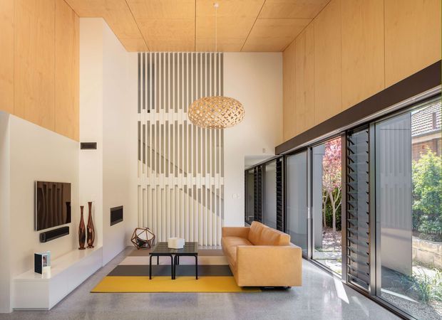 Bright contemporary living room with a double-height ceiling and extensive full-height glazing along one side. Warm timber wall and ceiling panels frame a white feature wall with vertical decorative battens, while a tan leather sofa, black coffee table, yellow area rug, sculptural pendant light, and minimalist media joinery create a refined modern interior. The glazing opens toward a landscaped exterior courtyard or garden.