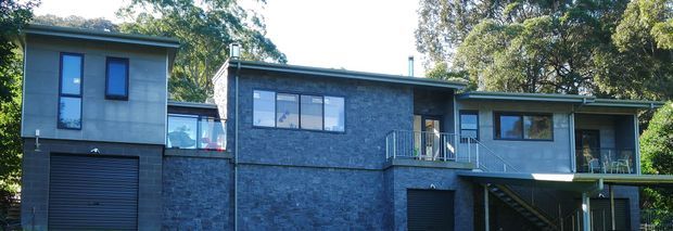A contemporary multi-level residential house set among mature trees, with dark grey stone or masonry cladding, grey upper-level walls, low-pitched rooflines, large rectangular windows, balconies, and external stairs. The facade includes an integrated garage and elevated sections stepping across the site.