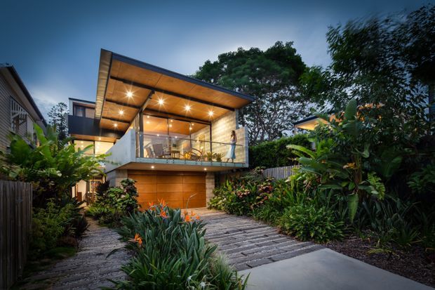 Twilight exterior view of a contemporary multi-level house set within lush tropical landscaping. The home features a dramatic projecting flat roof with exposed timber soffit, extensive upper-level glazing and a glass balustrade, warm timber cladding, concrete walls, and an integrated garage or entry level. A planted garden and textured paved driveway lead toward the building.