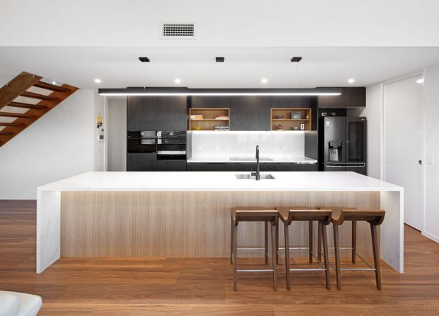 Contemporary open-plan kitchen dominated by a large white island with an integrated sink and three timber bar stools. The kitchen features dark wood cabinetry, black appliances, illuminated open shelving, a white splashback, and warm timber flooring. A timber staircase is visible at the left, while white walls and doors create a clean, modern backdrop.
