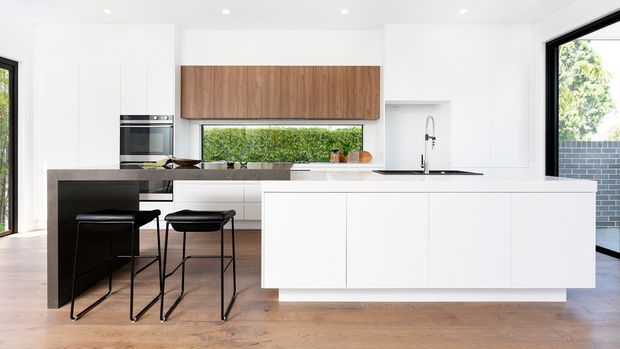 Bright contemporary kitchen with a large white island, integrated black sink and tap, dark stone benchtop, timber cabinetry, white walls and cabinetry, and timber flooring. Two black bar stools sit at the island, with large windows providing abundant natural light and views to greenery and a brick wall.