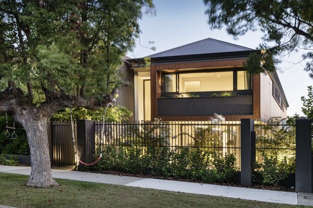 Exterior view of a contemporary two-storey residence behind a black metal fence, with a dark low-pitched roof, warm timber-framed upper-level glazing, pale rendered walls, and landscaped frontage with mature trees.