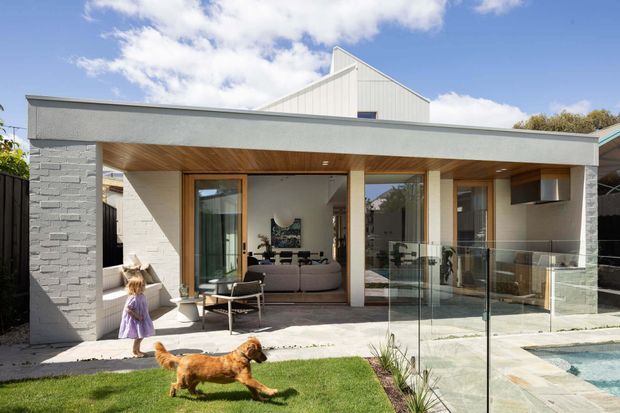 Modern single-storey residential extension with a low flat-roofed concrete or rendered wing, white textured masonry wall, timber-lined covered outdoor living area, extensive glazing, and a raised pitched-roof volume behind. The image shows a lawn, child and dog in the foreground, and a glass balustrade beside a small pool or water feature. Through the glazing, a contemporary living room is visible.