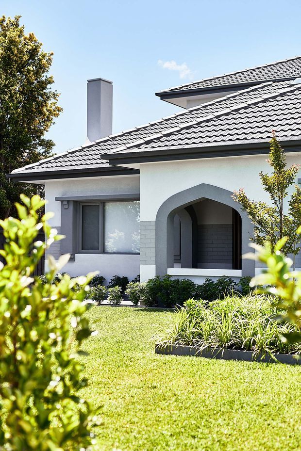 Exterior view of a light-coloured single-storey suburban house with a tiled pitched roof, prominent arched porch opening, chimney, rendered walls, and a neatly landscaped front garden with lawn and planted beds.
