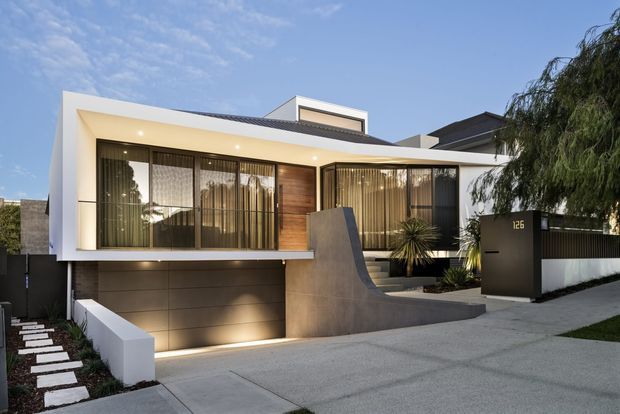 Street-facing view of a contemporary multi-level residence with a crisp white projecting upper facade, extensive floor-to-ceiling glazing, warm timber wall cladding, dark metal detailing, and a recessed garage. A sculptural concrete entry wall, landscaped front garden, paved driveway, and freestanding dark mailbox define the approach.