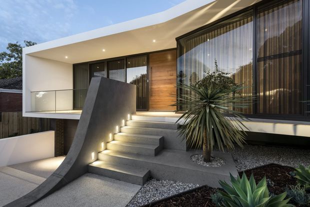 Contemporary residential facade viewed from the front, featuring a curved white roof canopy, extensive dark-framed glazing with sheer curtains, warm timber wall cladding, a cantilevered upper level, and a sculptural concrete stairway with integrated step lighting. Gravel landscaping and architectural succulents frame the entrance.