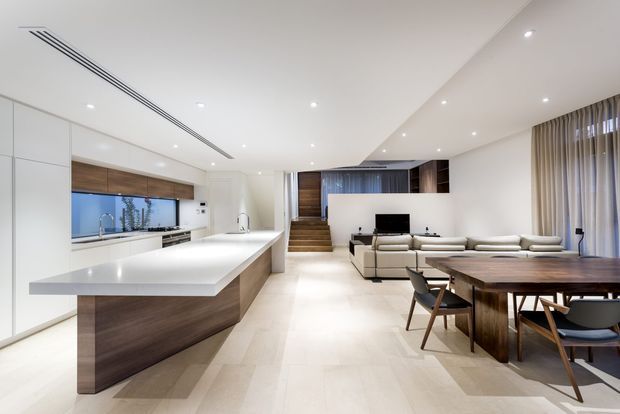 Open-plan contemporary interior combining a streamlined white kitchen with a large island, dining area, and adjacent living space. Warm timber cabinetry and flooring contrast with white surfaces, while recessed ceiling lighting, linear air vents, full-height curtains, and clean-lined furniture create a bright, refined atmosphere. A short staircase leads toward a darker timber-lined area at the rear.