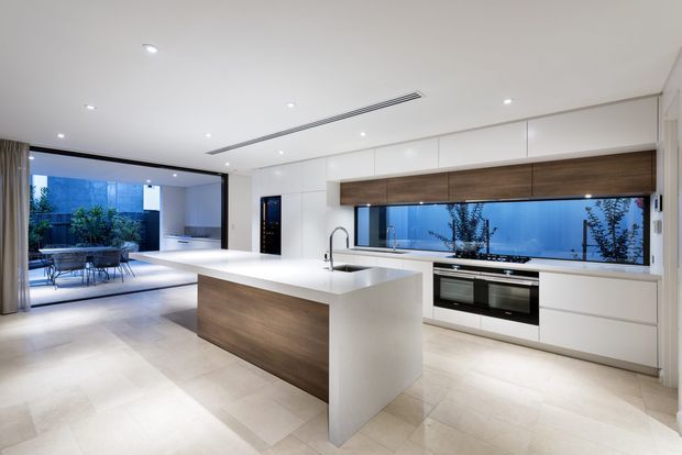 Contemporary open-plan kitchen with a large white island featuring a timber-panelled front, white full-height cabinetry, a long horizontal window, integrated appliances, and pale tiled flooring. The kitchen opens directly to a furnished outdoor terrace or courtyard through wide glazing, creating a bright, clean-lined indoor-outdoor connection.