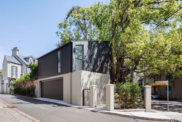Street-level view of a compact contemporary multi-storey urban residence with a dark vertically clad upper volume, pale rendered lower level, integrated garage, tall glazing, and a gated side entry. Mature street trees and neighbouring traditional homes frame the property.