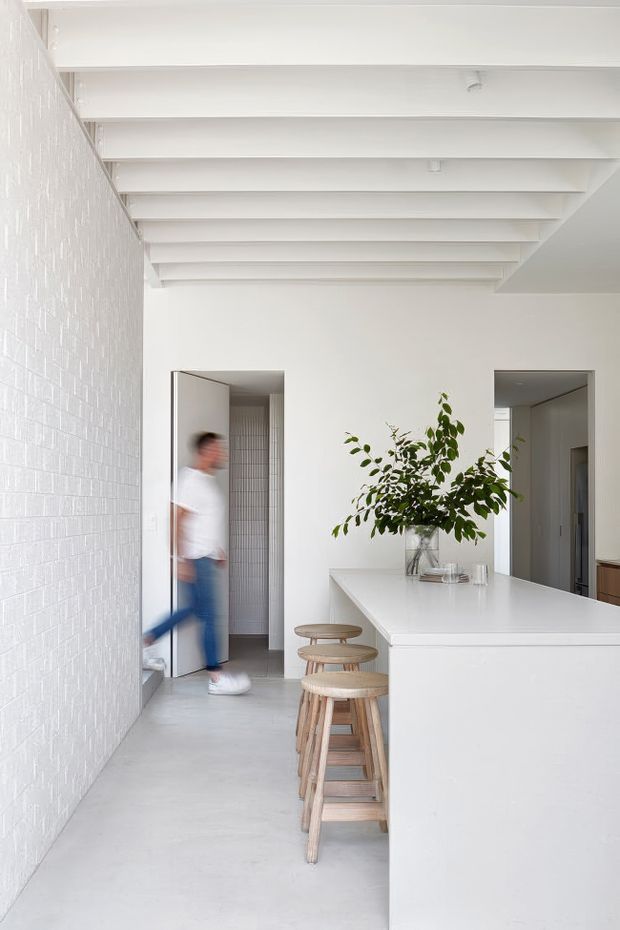Bright, minimalist kitchen interior with a large white island and integrated white cabinetry, paired with light timber stools. The space features a white textured brick wall, polished pale concrete flooring, exposed painted ceiling beams, and a simple leafy arrangement on the island. Doorways lead to adjoining areas in the background.
