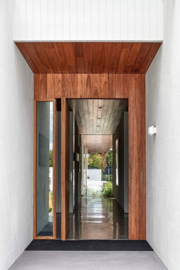 A refined residential entry viewed from outside, featuring a tall timber-framed doorway with a glazed side panel, rich dark wood lining and a dramatic timber soffit. The entrance opens into a long, polished hallway with dark walls, timber ceiling lining, reflective flooring, and a landscaped garden visible at the far end. White textured masonry walls frame the entry.
