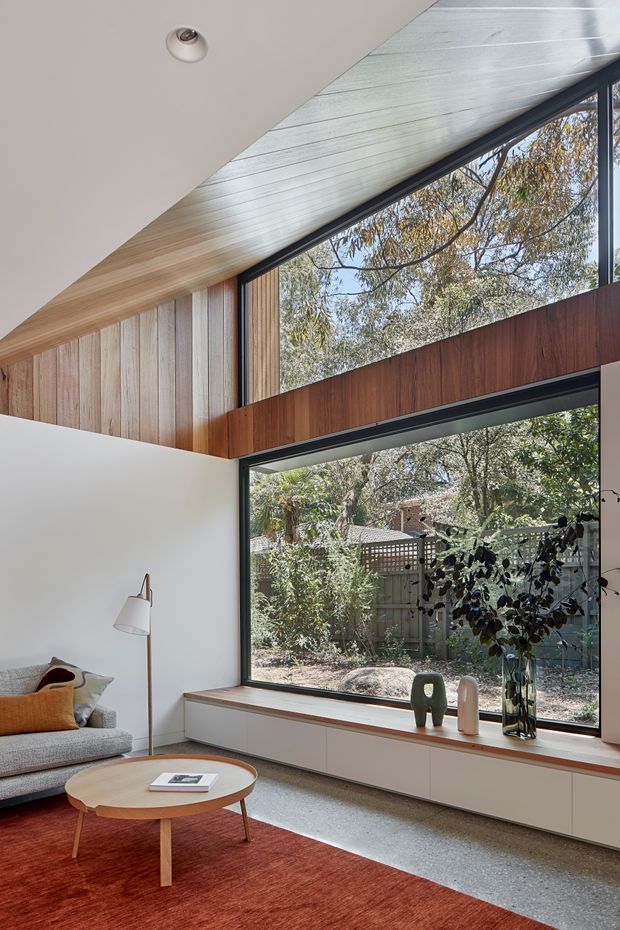 A bright, refined living room with a large corner window and clerestory glazing overlooking a leafy garden. The space features white walls, extensive vertical timber wall panelling, a sloping timber-lined ceiling, dark window frames, a built-in timber-topped window seat with white cabinetry, a grey sofa, a round timber coffee table, and a rust-red area rug. Natural light and mature trees create a calm indoor-outdoor connection.