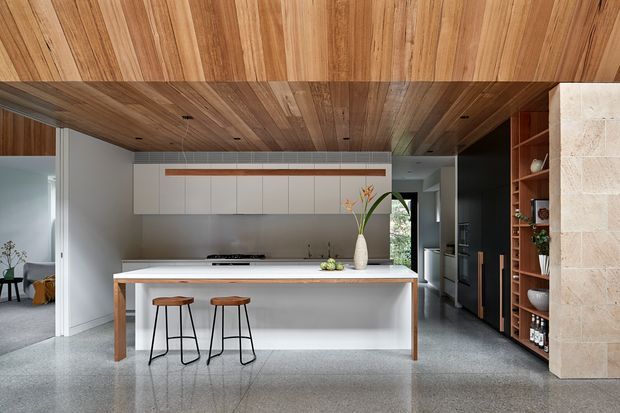 Contemporary open-plan kitchen with a large white island and timber legs, two stools, sleek white wall cabinetry, integrated appliances, and a warm timber-lined ceiling. A built-in timber display unit and pale stone wall add texture, while polished grey flooring and a view toward adjoining living areas complete the space.