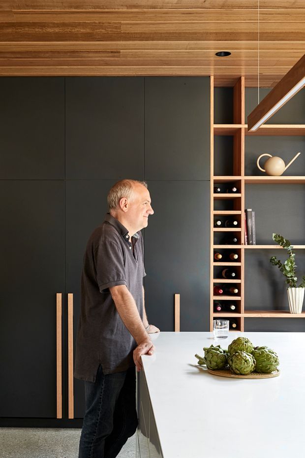 A refined contemporary kitchen interior with a large white island in the foreground, dark matte full-height cabinetry with long timber handles, and open timber shelving incorporating a narrow wine rack and decorative niches. The ceiling is clad in warm timber boards, complemented by timber shelving and structural detailing. A person stands beside the island, which is styled with artichokes and a glass of water. The palette is warm timber, charcoal, white, and muted grey, creating a sophisticated, calm atmosphere.